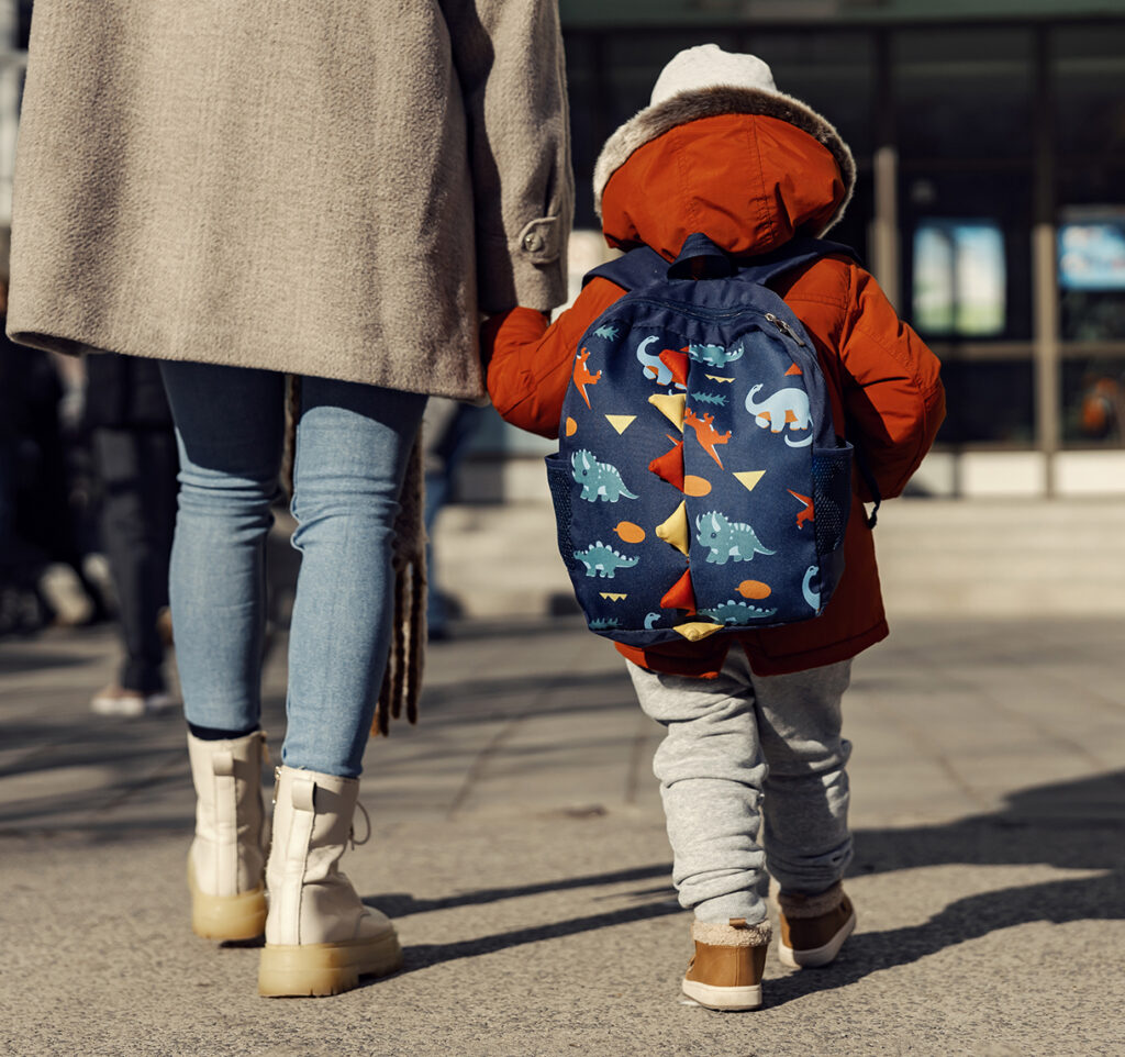 Rearview of mother walking preschooler into child care center or school setting with winter coats on; child has backpack hanging from shoulders.