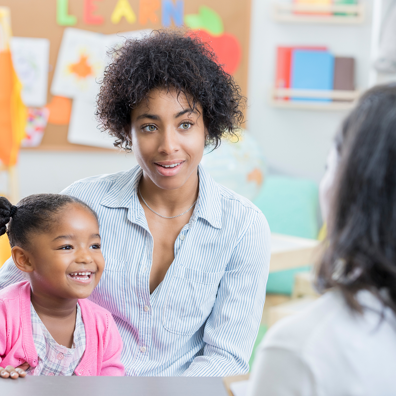 A young mother holds her preschool age daughter in her lap as she sits across a table from her daughter's new teacher for a meeting.