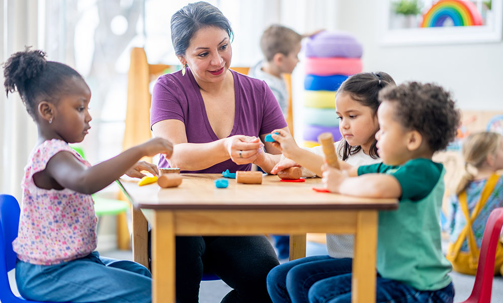 Preschool teacher sitting at table with 3 preschoolers playing with Play-Doh.