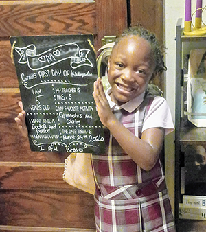 A five-year-old girl in school uniform smiling holding a chalk sign for her first day of kindergarten