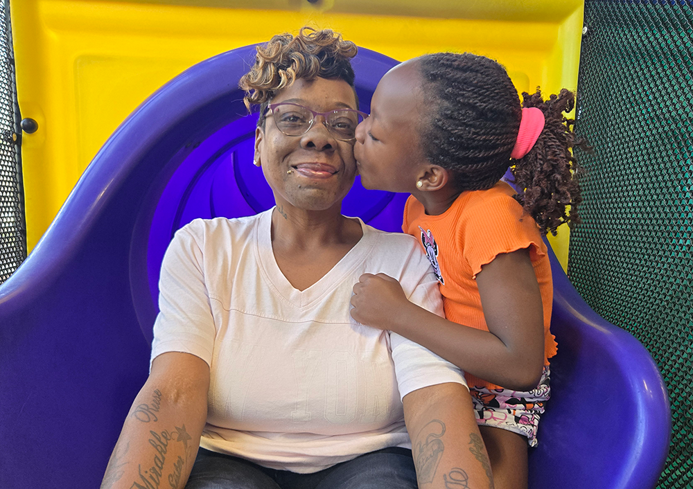 Black mother sitting at base of tube slide smiling as five-year-older daugher kisses her on the cheek.